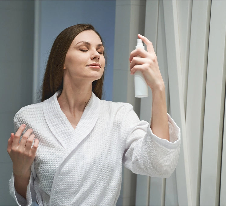Woman in a white robe applying a product from a spray bottle against a neutral background - e11ement