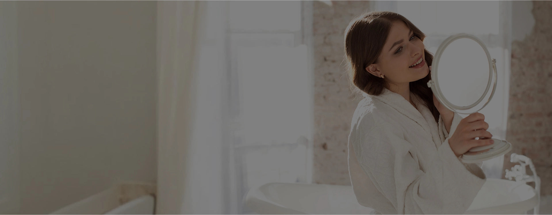 Woman in a white robe sitting by a bathtub holding a round mirror. e11ement